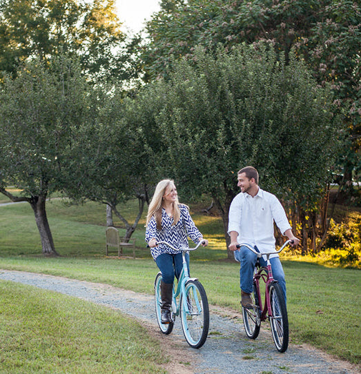 Two people ride bicycles side by side on a winding path through a green park, surrounded by trees and grass. They are smiling and looking at each other on a sunny day.