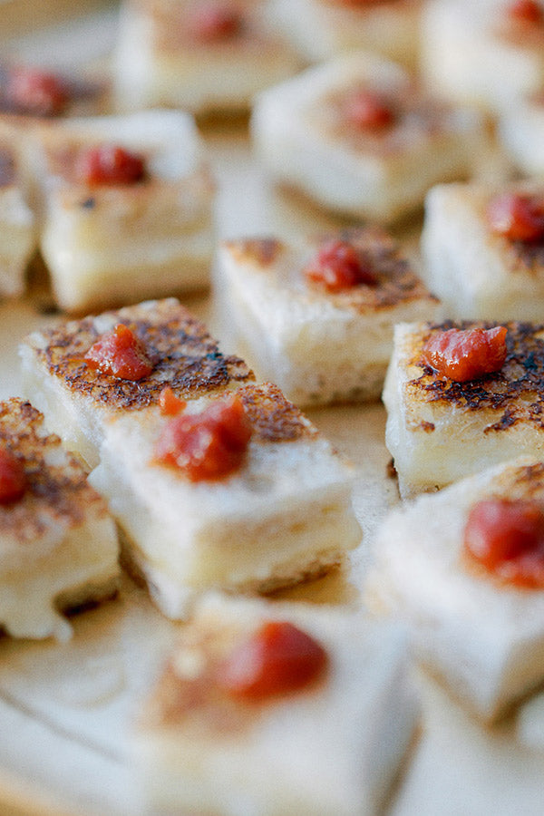 Close-up of several square pieces of grilled food, each topped with a small dollop of red sauce, arranged on a light-colored surface.