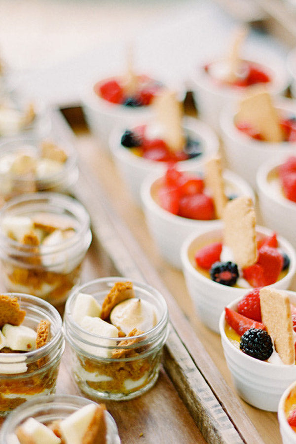 Rows of small dessert cups filled with layered fruit, cream, and cookies or biscuits, placed on a wooden tray. Some cups contain berries and sliced strawberries, while others have bananas and crumbled cookies.