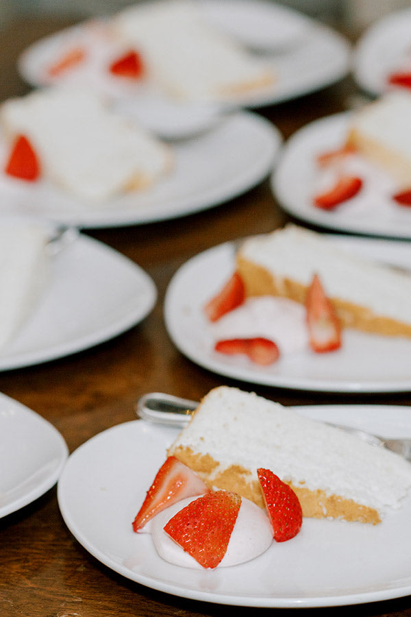 Several white plates each hold a slice of white frosted cake garnished with fresh strawberry pieces and a dollop of whipped cream, arranged on a wooden surface.