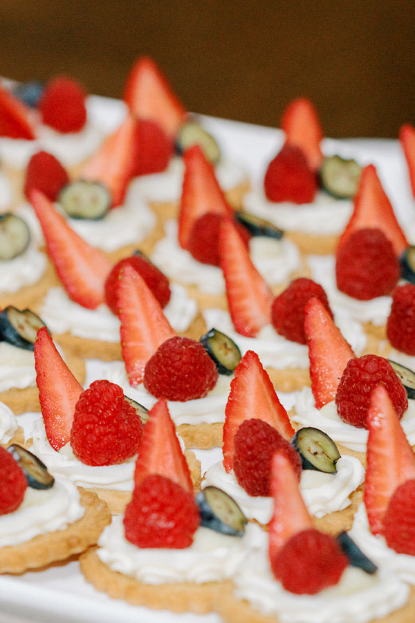 Assorted mini tarts topped with whipped cream, halved strawberries, raspberries, and sliced blueberries are arranged on a white serving tray.