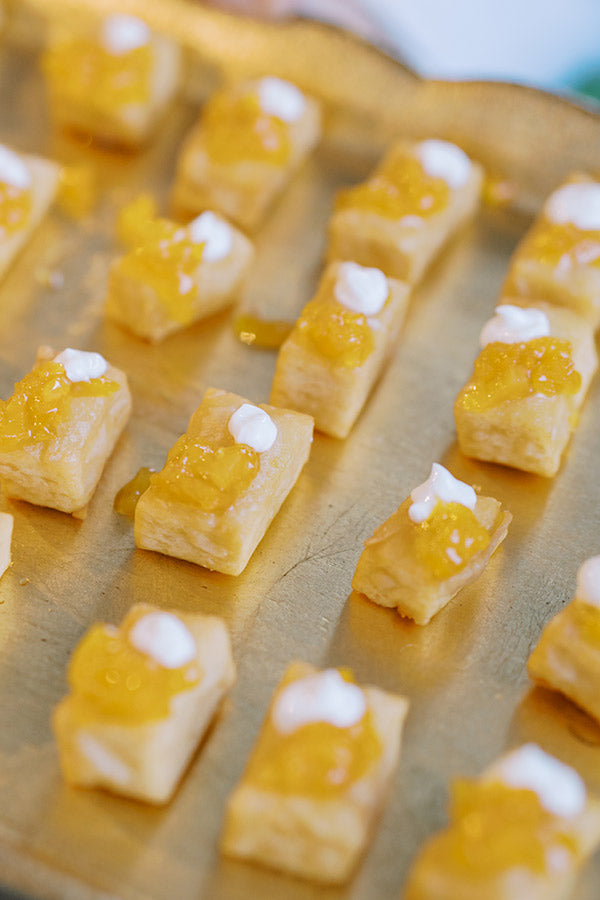 Small rectangular pastries topped with yellow fruit preserves and a dollop of white cream are arranged in neat rows on a gold tray.