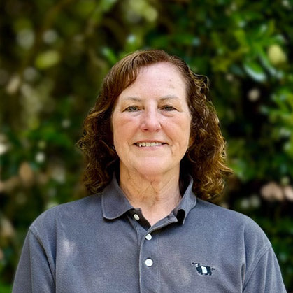 A middle-aged woman with curly brown hair smiles while standing outdoors in front of green foliage. She is wearing a gray collared shirt with a small logo on the chest.