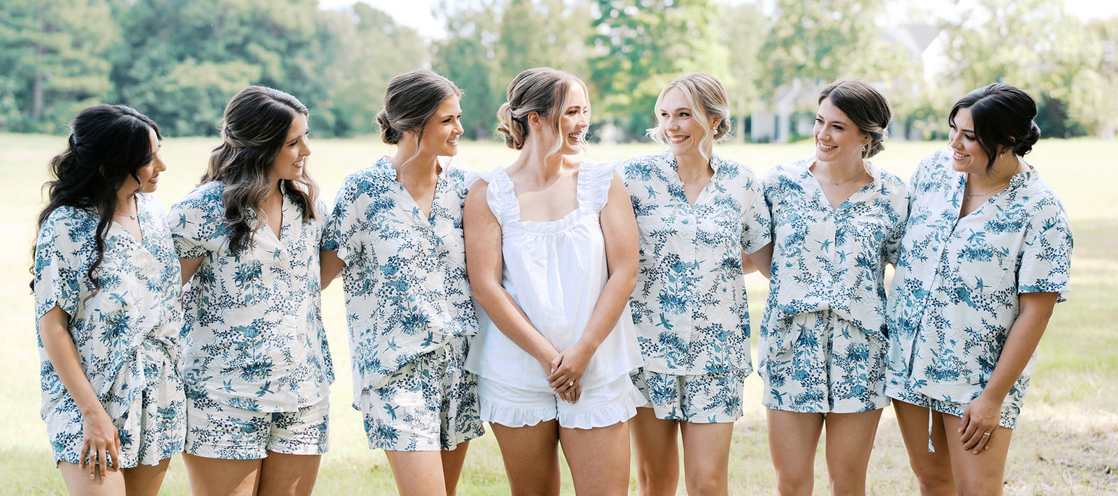 Seven women stand outdoors, smiling at each other. Six wear matching blue and white floral pajamas, while the woman in the center wears a white ruffled pajama set. The background shows trees and grass.