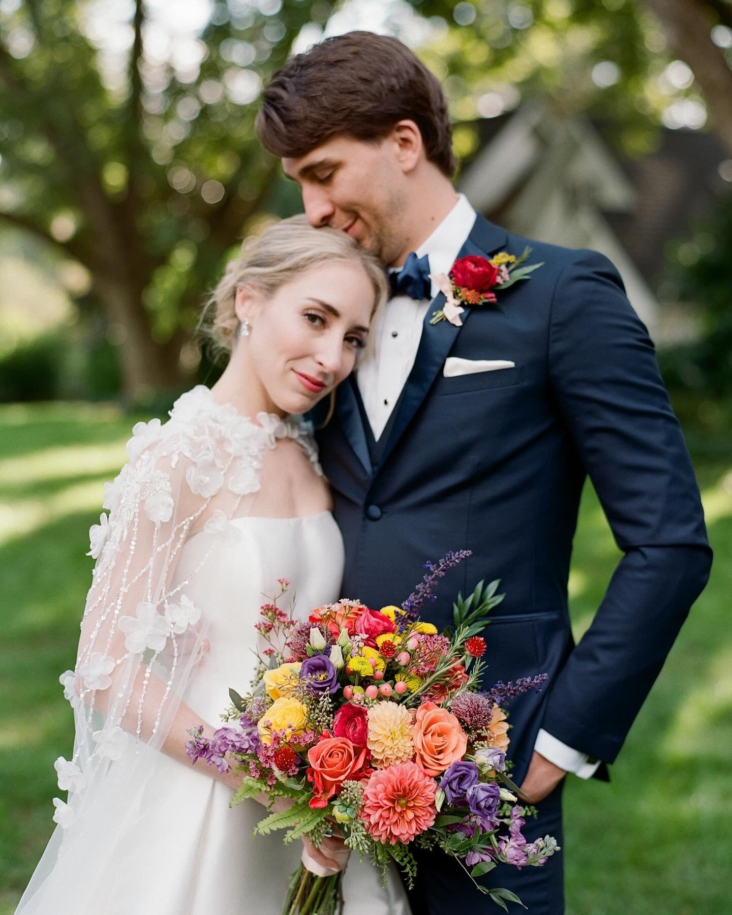 A bride in a white dress with a floral cape leans her head on a groom’s chest outdoors. The groom wears a navy suit with a boutonniere. The bride holds a colorful bouquet. Both appear happy and serene.