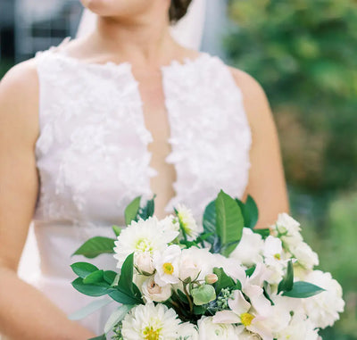 A bride in a sleeveless white lace gown holds a bouquet of white flowers and greenery. Her face is turned to the side, and the background is blurred with greenery.