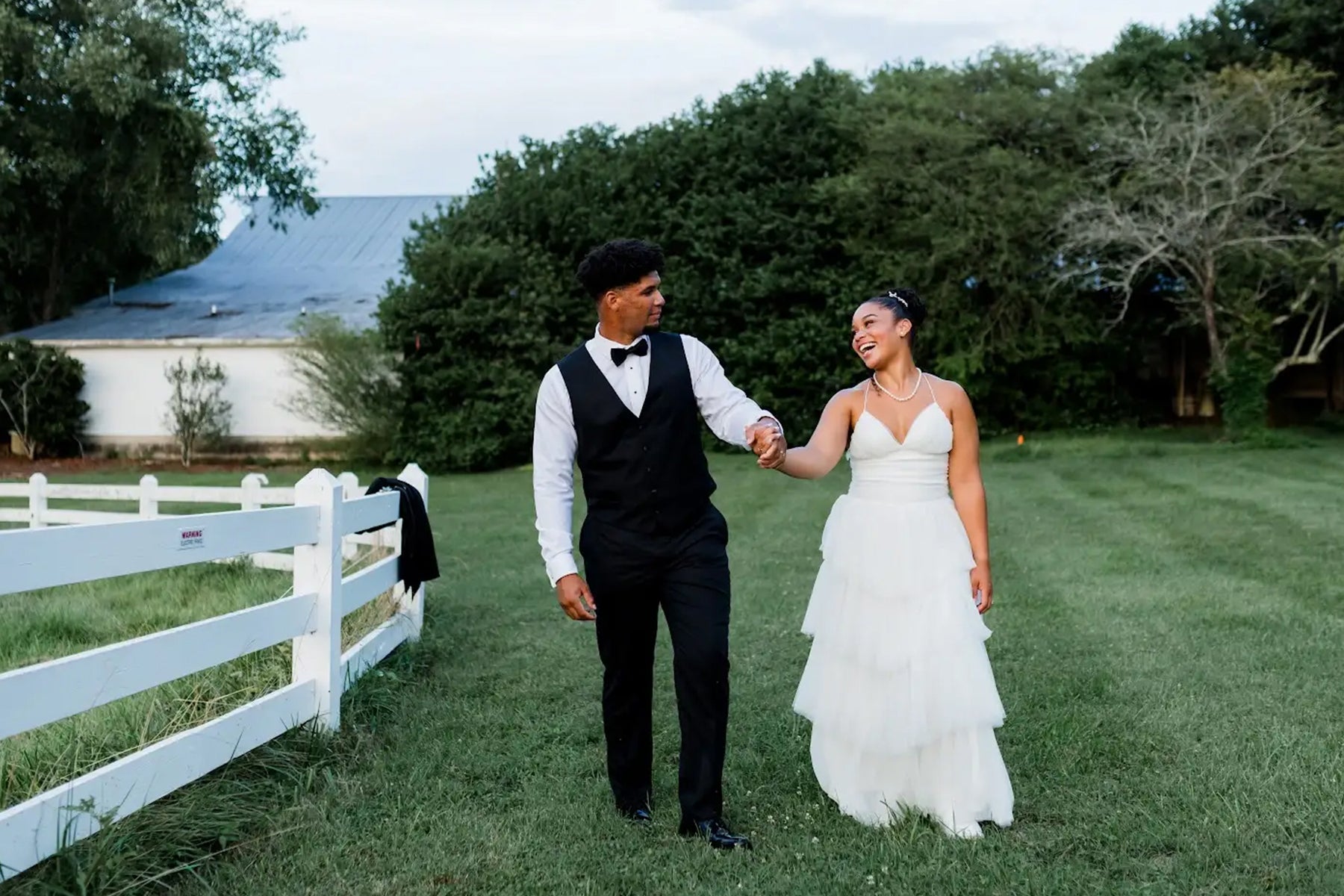 A couple dressed in wedding attire walk hand in hand on grass, smiling at each other. The groom wears a black vest and bow tie, while the bride wears a white gown. Trees and a white fence are in the background.