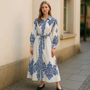 A woman with light brown hair stands on a paved sidewalk in front of a cream-colored building, wearing the FARYLINA SHERI LONG SLEEVE DRESS by FARYLINA—white cotton with blue florals, indigo embroidery, and a belted waist—plus black shoes.