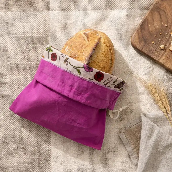 A round loaf of bread in the BEES WRAP - BEESWAX REUSABLE BREAD BAKERY BAG MAGENTA by BEES WRAP, featuring a magenta cloth and floral trim, is placed on a textured tablecloth beside a wooden cutting board, wheat stalks, and a folded napkin.
