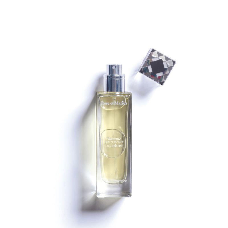 A rectangular glass ROSE ET MARIUS perfume bottle with a silver spray nozzle and diamond-patterned cap sits beside it, labeled &quot;THE MAN WHO PLANTED TREES&quot; Eau de Parfum 30ml, displayed on a white background.