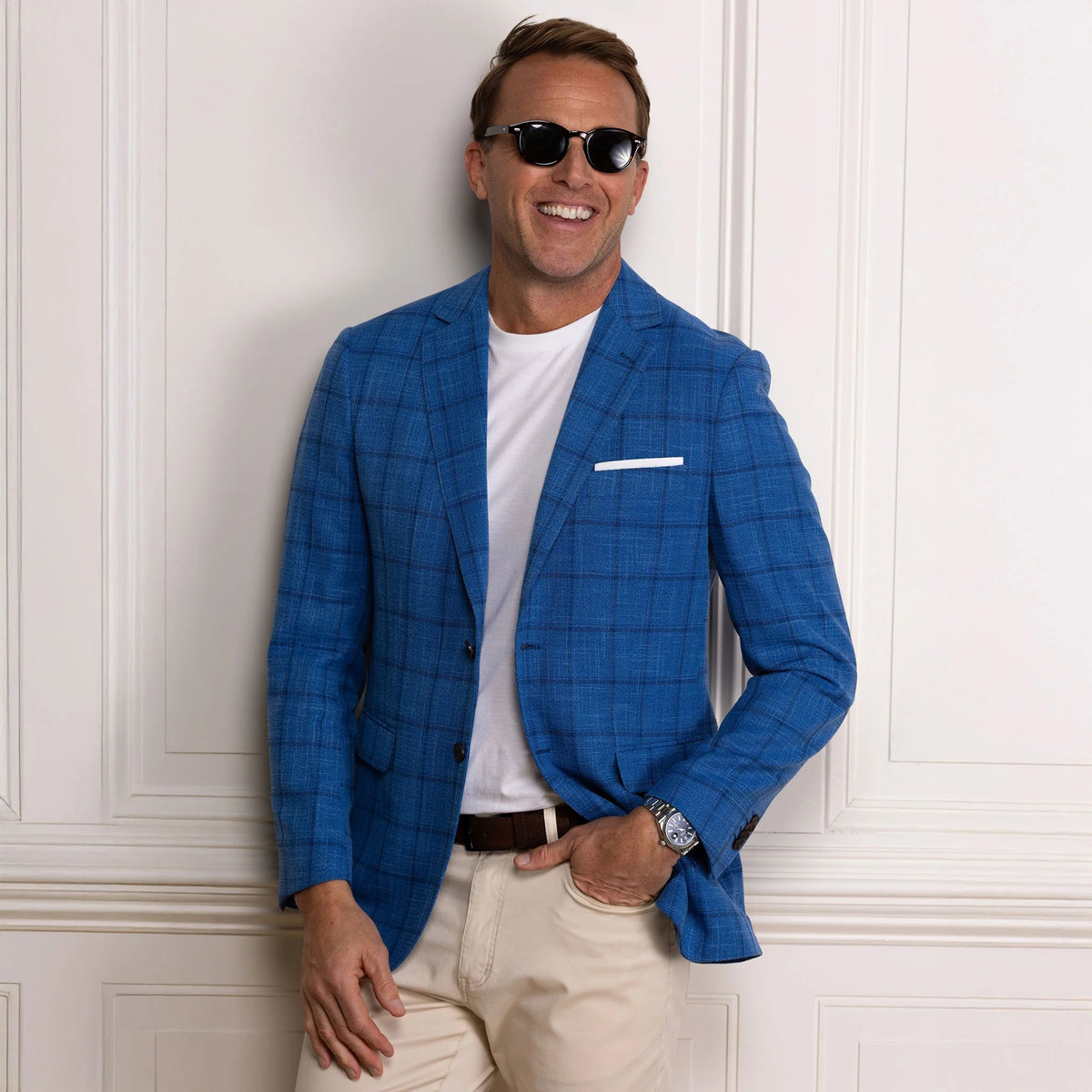 A man wearing sunglasses, the COLLARS AND CO MAVERICK TRAVEL BLAZER IN BLUE WINDOWPANE over a white t-shirt, along with light pants and a brown belt, stands smiling with one hand in his pocket against a white paneled wall.