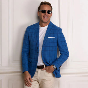 A man wearing sunglasses, the COLLARS AND CO MAVERICK TRAVEL BLAZER IN BLUE WINDOWPANE over a white t-shirt, along with light pants and a brown belt, stands smiling with one hand in his pocket against a white paneled wall.