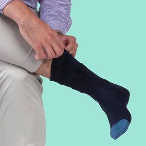 A person in light pants and a purple checkered shirt pulls up BIRD DOG BAY - PEDIGREE MID-CALF SOCKS IN GREEN by BIRD DOG BAY against a light teal background.