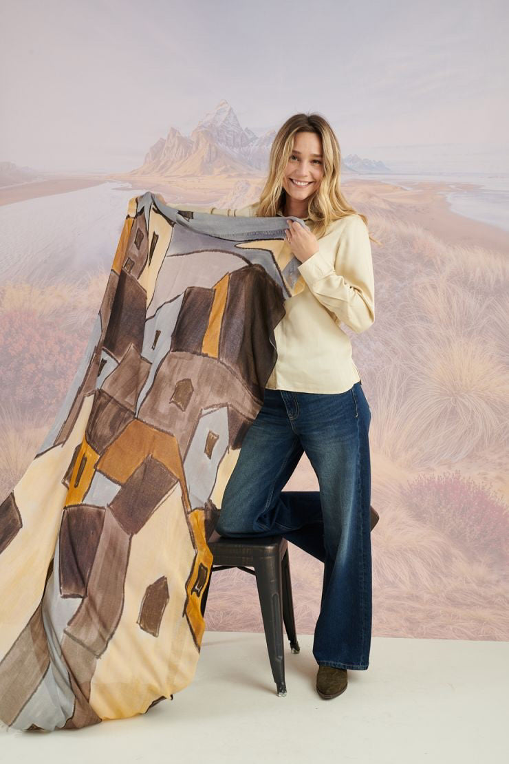 A woman in a black long-sleeve top and beige pants stands before a scenic backdrop, her look elevated by the STORIATIPIC ISCHIA Modal and Wool Scarf (100x200 cm) in vibrant orange and brown, draped elegantly around her neck.