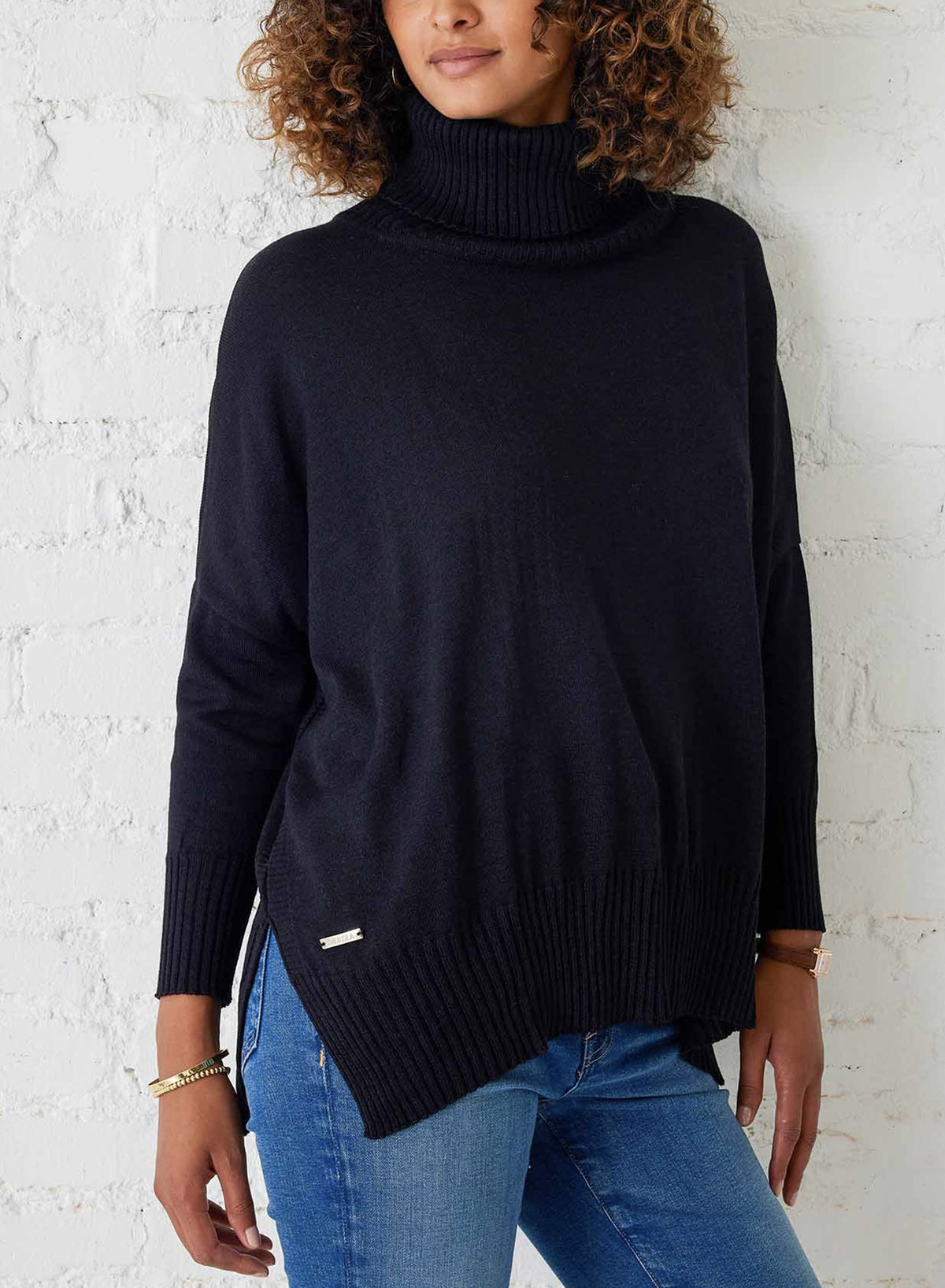 A woman in the MERSEA Catalina Turtleneck Sweater in True Black and blue jeans stands by a white brick wall, her curly hair styled naturally and gold bracelets adorning her wrist.