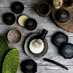 A round puck of THE IMMACULATE BEARD - SHAVE SOAP in First Light, ideal for sensitive skin, rests in a gray bowl. The black label features sandalwood, vanilla, and other notes from THE IMMACULATE BEARD.