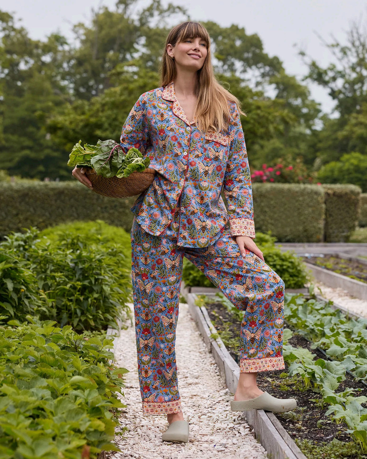 A woman with light brown hair sits against a white wall, wearing the PRINTFRESH - Bee Tapestry Long Pajama Set in Delphinium Blue. This organic cotton pajama set from PRINTFRESH features a colorful floral and bee pattern with vibrant red and yellow cuffs, collar, and pocket trim, beautifully blending playful design with cozy comfort.