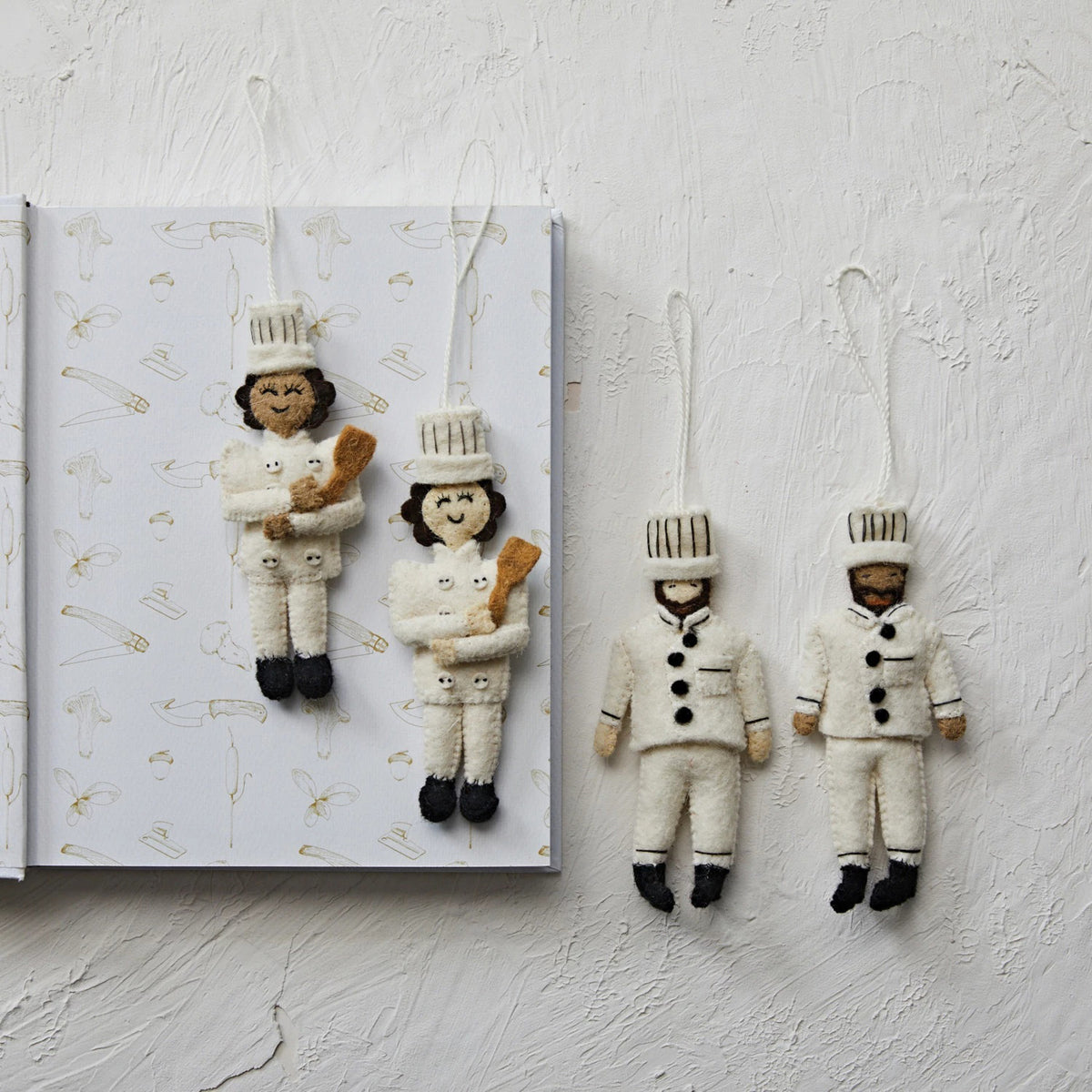 Four CREATIVE COOP FELT CHEF ORNAMENTS—two hanging from a festive book, two on a white textured surface. Each handmade, embroidered chef wears a white uniform and hat; two hold brown baguettes.