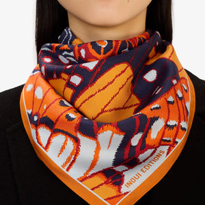A person in a dark grey knit sweater wears an INOUI EDITIONS PAPILLONS Orange Silk Scarf, featuring bold abstract butterfly designs and “INOUI EDITIONS” on the border, neatly tied around their neck.