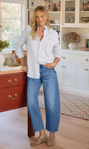 A woman smiles in a bright kitchen, wearing the FRANK & EILEEN EILEEN Relaxed Button Up Sweatshirt in Grey Melange. She pairs it with blue wide-leg jeans and tan boat shoes, leaning against a red table with one hand in her pocket.