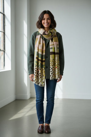 A woman stands indoors, smiling, wearing a green coat, blue jeans, brown shoes, and the STORIATIPIC - BAO Wool Scarf with Embroidery in Khaki by STORIATIPIC. Sunlight streams through a window on the left, illuminating the room’s white walls and light floor.