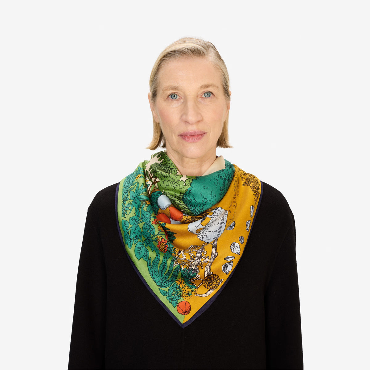 A woman wears the INOUI EDITIONS - QUATRE SAISON MODAL SILK SCARF by INOUI, featuring vibrant green, yellow, and orange patterns of trees, flowers, and abstract designs. Only her lower face, neck, and upper chest are visible. Made in India.