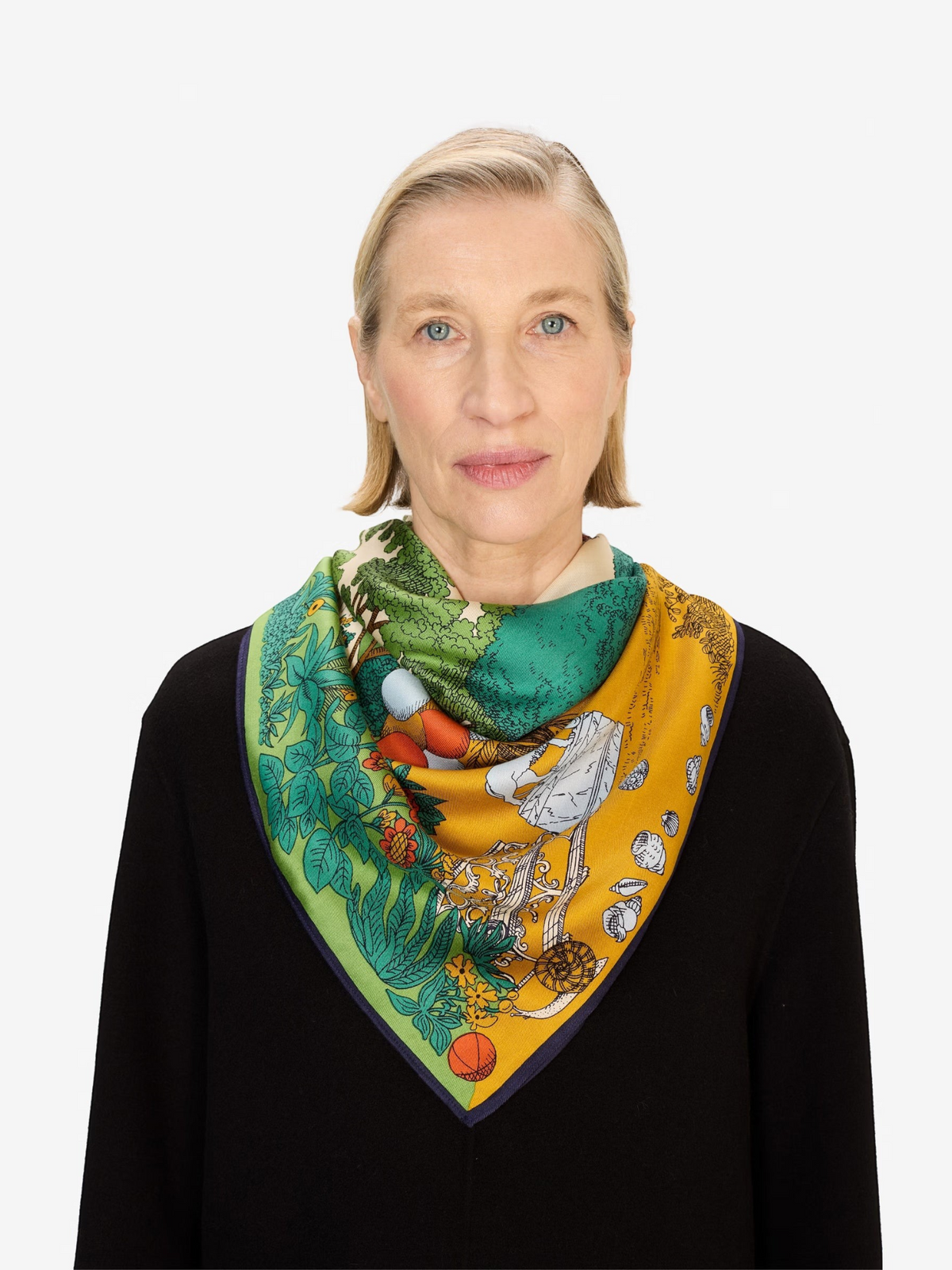 A woman wears the INOUI EDITIONS - QUATRE SAISON MODAL SILK SCARF by INOUI, featuring vibrant green, yellow, and orange patterns of trees, flowers, and abstract designs. Only her lower face, neck, and upper chest are visible. Made in India.