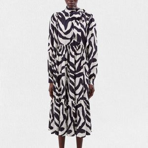 A person is wearing the MEIMEIJ Ruffle Print Dress, a long-sleeved, black and white zebra print midi dress made from 100% viscose fabric. It features a high neckline, gathered waist, tiered skirt, and buttoned cuffs. The background is plain white.