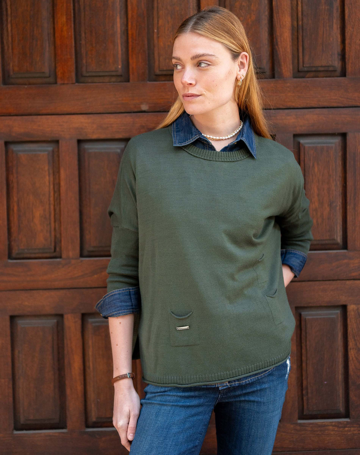 A woman with light brown hair stands before a wooden door, wearing the MERSEA - CATALINA CREWNECK SWEATER IN DEEP EMERALD over a denim shirt and blue jeans, gazing to the side with hands in her pockets and a relaxed expression.