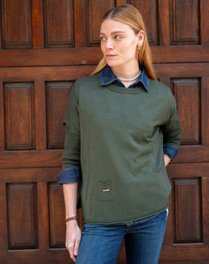 A woman with light brown hair stands before a wooden door, wearing the MERSEA - CATALINA CREWNECK SWEATER IN DEEP EMERALD over a denim shirt and blue jeans, gazing to the side with hands in her pockets and a relaxed expression.