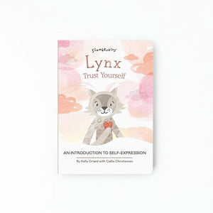 A small child’s hand points to an open SLUMBERKINS - LYNX TRUST YOURSELF BOOK. The left page contains text about feelings and how to be brave and speak up. The right page features an illustration of a smiling cat with a heart on its chest, sitting cross-legged in a peaceful garden with flowers and clouds.