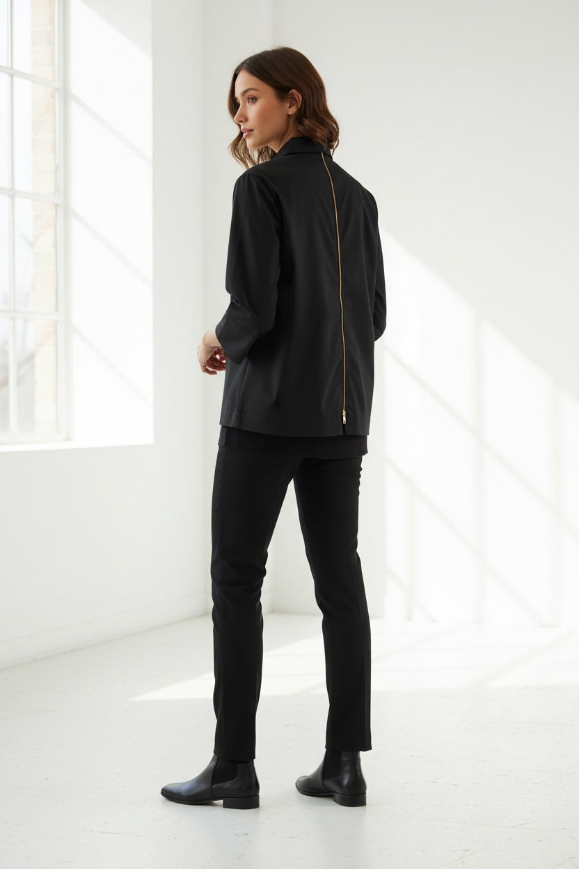 Standing against a light-colored wall with intricate molding, a woman with long hair reminiscent of Xena is turned away from the camera, showcasing the HINSON WU - XENA ZIP BACK SHIRT in navy blue. This A-line shaped top features an eye-catching gold tone zipper that extends down her back, perfectly complemented by elegant black pants.