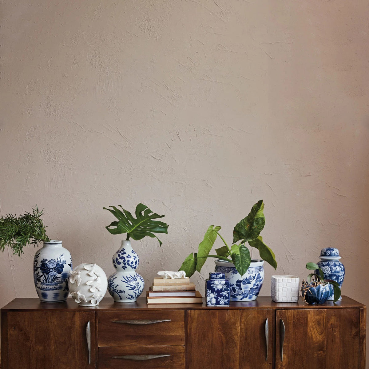 A wooden cabinet is adorned with VASE WITH BIRDS by CREATIVE COOP, handmade stoneware pieces showcasing intricate blue and white designs, complemented by assorted potted plants. The setup stands against a plain beige wall, enhancing the minimalist elegance of the decor.