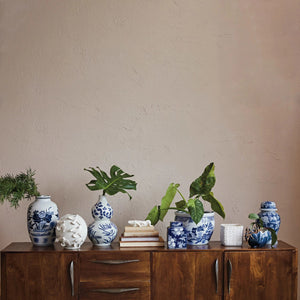 A wooden cabinet is adorned with VASE WITH BIRDS by CREATIVE COOP, handmade stoneware pieces showcasing intricate blue and white designs, complemented by assorted potted plants. The setup stands against a plain beige wall, enhancing the minimalist elegance of the decor.