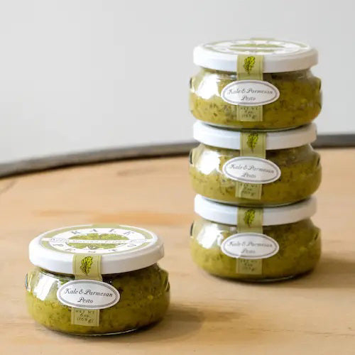 A small, round jar with a white lid labeled "BELLA CUCINA - KALE &amp; PARMESAN PESTO." The jar is filled with green, all-natural pesto sauce and features a decorative label displaying the product name and net weight of 6 ounces (169 grams).