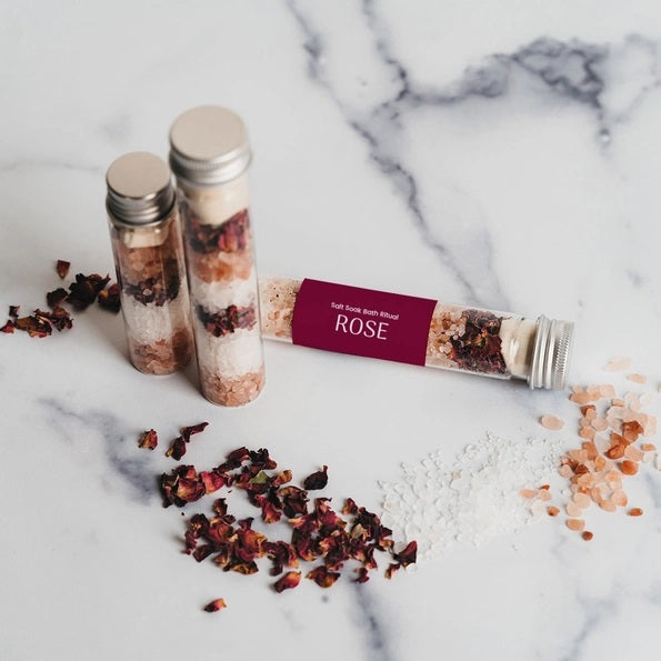 Three BOTANICAL BATH SALT TUBES - ROSE by GIFTEN MARKET filled with layered pink and white rose bath salts and dried rose petals are arranged on a marble surface, with some of the mineral-rich salts and petals scattered around.