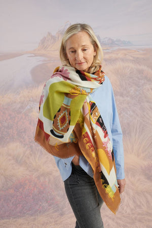 A woman with light brown hair wears a cream blouse and the STORIATIPIC - ANDRES WOOL SCARF by STORIATIPIC, elegantly hand-embroidered with green, yellow, red, and blue patterns. She gazes to the side against a scenic background.