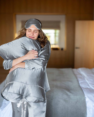 A woman in MERSEA’s Over The Moon Satin Sailor Sleep Mask sits on a bed, smiling gently as she lifts the mask from her eyes, with a softly blurred, lavender-toned background enhancing the calm mood.