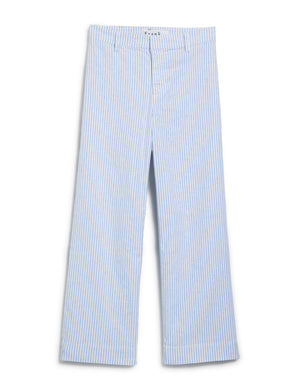 Standing on a stone surface, a person wears a denim shirt with FRANK & EILEEN Wexford Wide Leg Pant in blue and white stripe, complemented by sandals. They are outdoors amid lush greenery with arms relaxed by their sides.