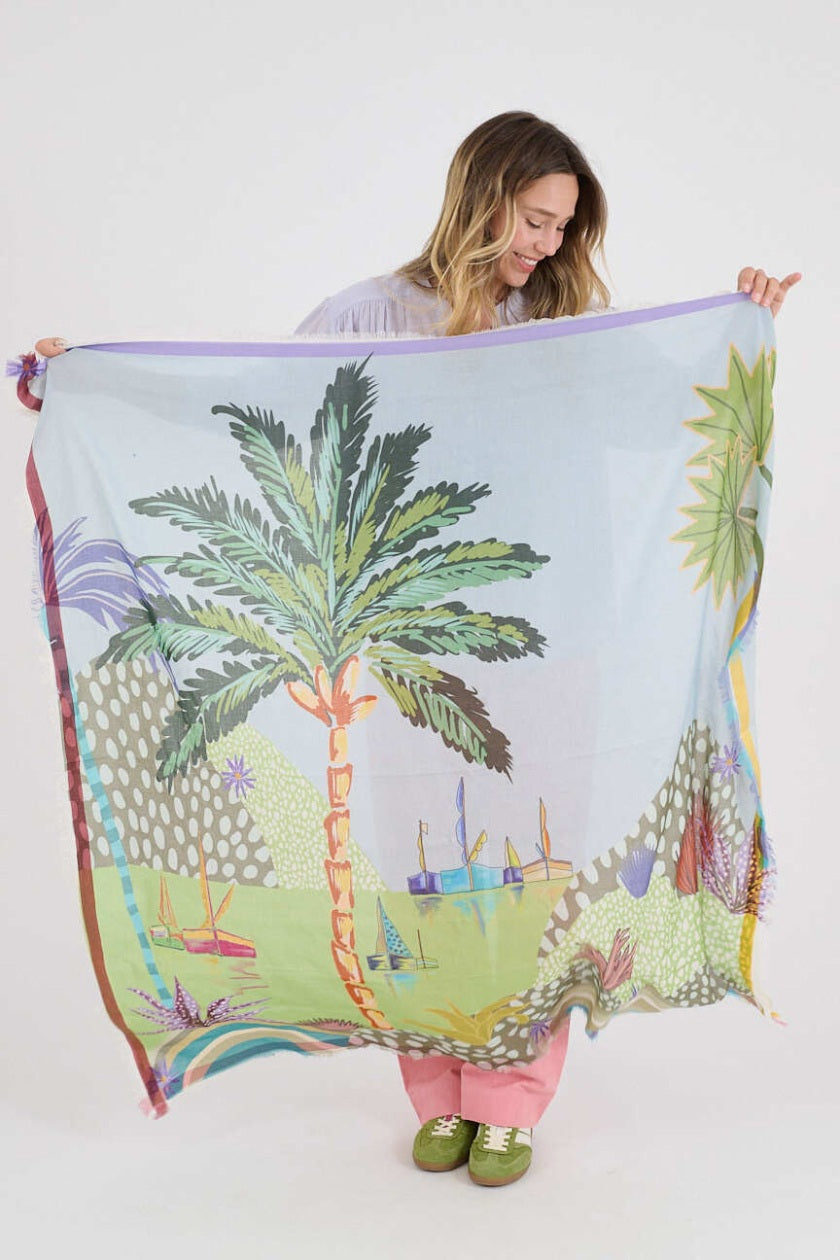 A woman with light brown hair wears a pale purple button-up shirt, pink pants, and the STORIATIPIC - CALI MAX SCARF—a large, colorful tropical-print accessory by STORIATIPIC—draped around her shoulders against a plain white background.