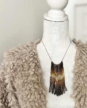 A mannequin wears a textured, curly taupe jacket and the ANN LIGHTFOOT Tiger Eye and Quartz Ombre Cascade Necklace, featuring dramatic dark beaded fringes in a triangular shape. The softly blurred background is neutral.