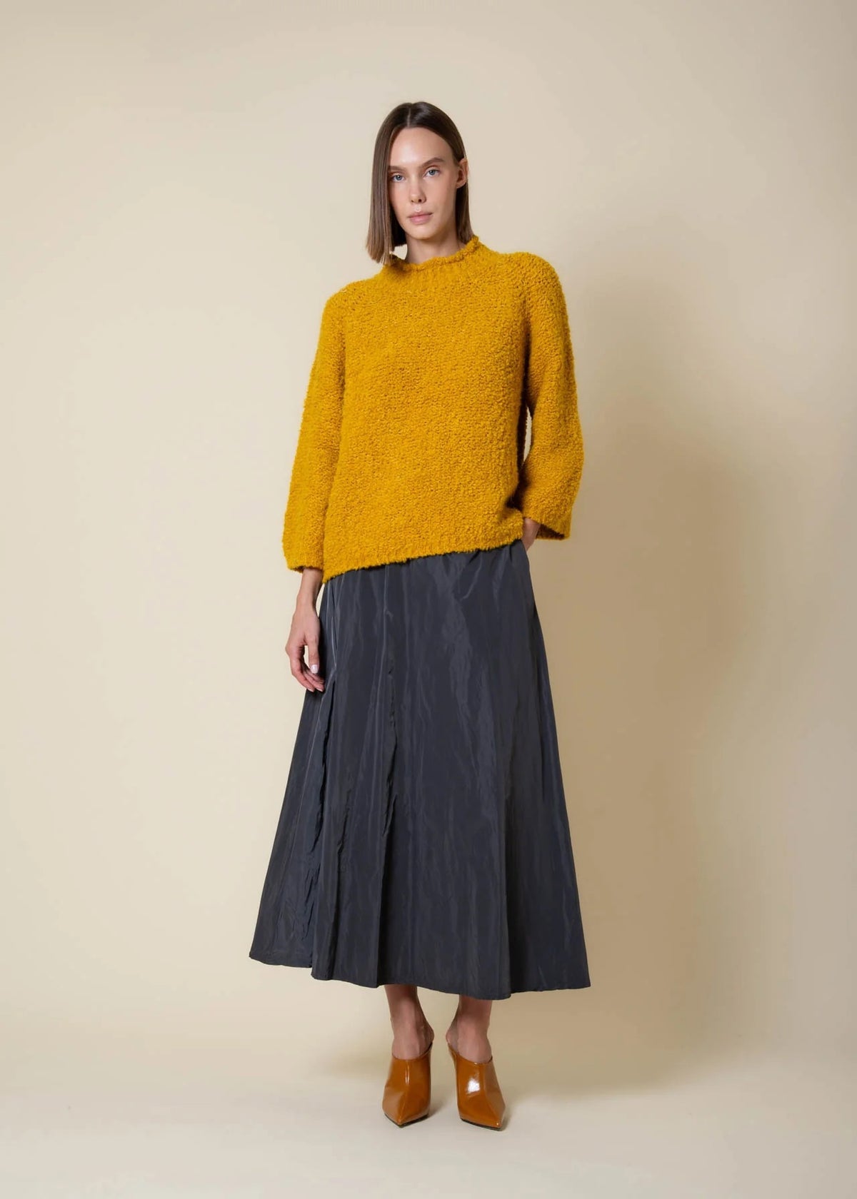 Wearing the VICARIO CINQUE - MADEIRA Bouclé Sweater in Mustard by VICARIO CINQUE, a woman pairs it with a long black pleated skirt and brown pointed-toe shoes, standing against a beige background.