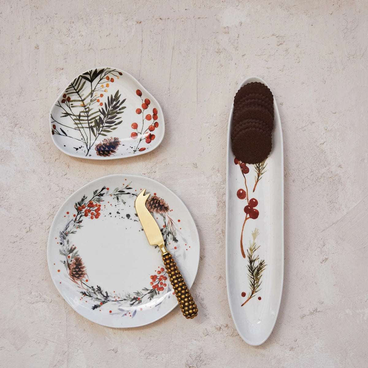 Three CREATIVE COOP STONEWARE PLATES WITH SEASONAL BOTANICALS—a round plate with a gold cheese knife, a triangular plate, and a long oval plate with cookies—display berry and floral designs on a light textured surface.