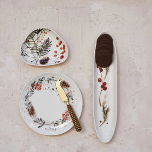 Three CREATIVE COOP STONEWARE PLATES WITH SEASONAL BOTANICALS—a round plate with a gold cheese knife, a triangular plate, and a long oval plate with cookies—display berry and floral designs on a light textured surface.