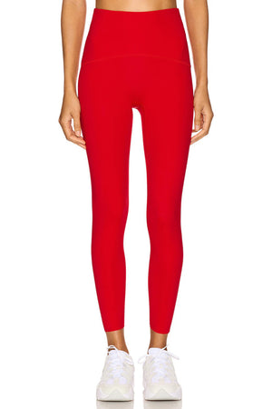 A person wears SPANX - BOOTY BOOST 7/8 LEGGINGS in high-waisted royal blue performance fabric with black athletic shoes, standing hand on hip against a plain white background. Only their lower body is visible; upper body and face are not shown.