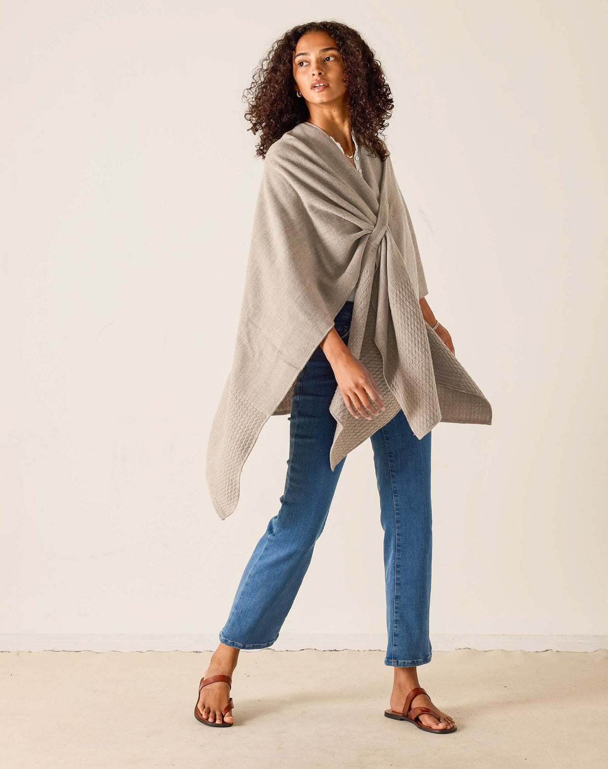 A woman stands indoors in a bright room with light streaming through a large window, smiling softly. She wears her natural curly hair loose and is styled in blue jeans, brown sandals, and a white shirt layered with the MERSEA - MODERN TRAVEL WRAP IN DRIFTWOOD by MERSEA, featuring a securing loop.