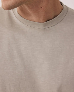 A man with short hair wears the HARTFORD Slub Cotton Crewneck T-shirt in Sand and green pants, standing with one hand in his pocket and looking to the side against a light background.