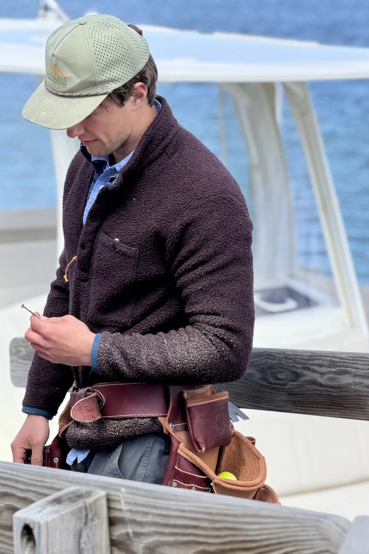 A man wearing a green cap, brown fleece jacket, and tool belt stands by a wooden railing, examining a nail in his hand. His rugged look is completed with the RELWEN - WOOLY SHAG SNAP-ZIP IN BLACKBERRY MARL layered beneath his jacket. A boat and water are visible in the background.