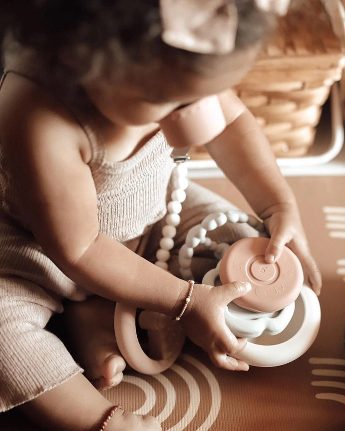 Introducing the SILICONE SUN STACKING RING TOWER from ALI + OLI: a set of BPA-free silicone stacking rings in soft pastel hues. The rings come in various shapes and textures, including hearts, and feature a sun-shaped topper on a sturdy base. This baby development toy is designed to promote motor skills and creativity.