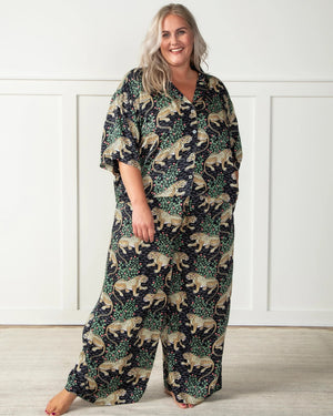 A woman stands barefoot on a woven rug in front of a white paneled wall, wearing the PRINTFRESH - Bagheera Satin Wildest Dream Pajama Set in Ink from PRINTFRESH, featuring a vibrant leopard and foliage pattern with wide-leg pants.